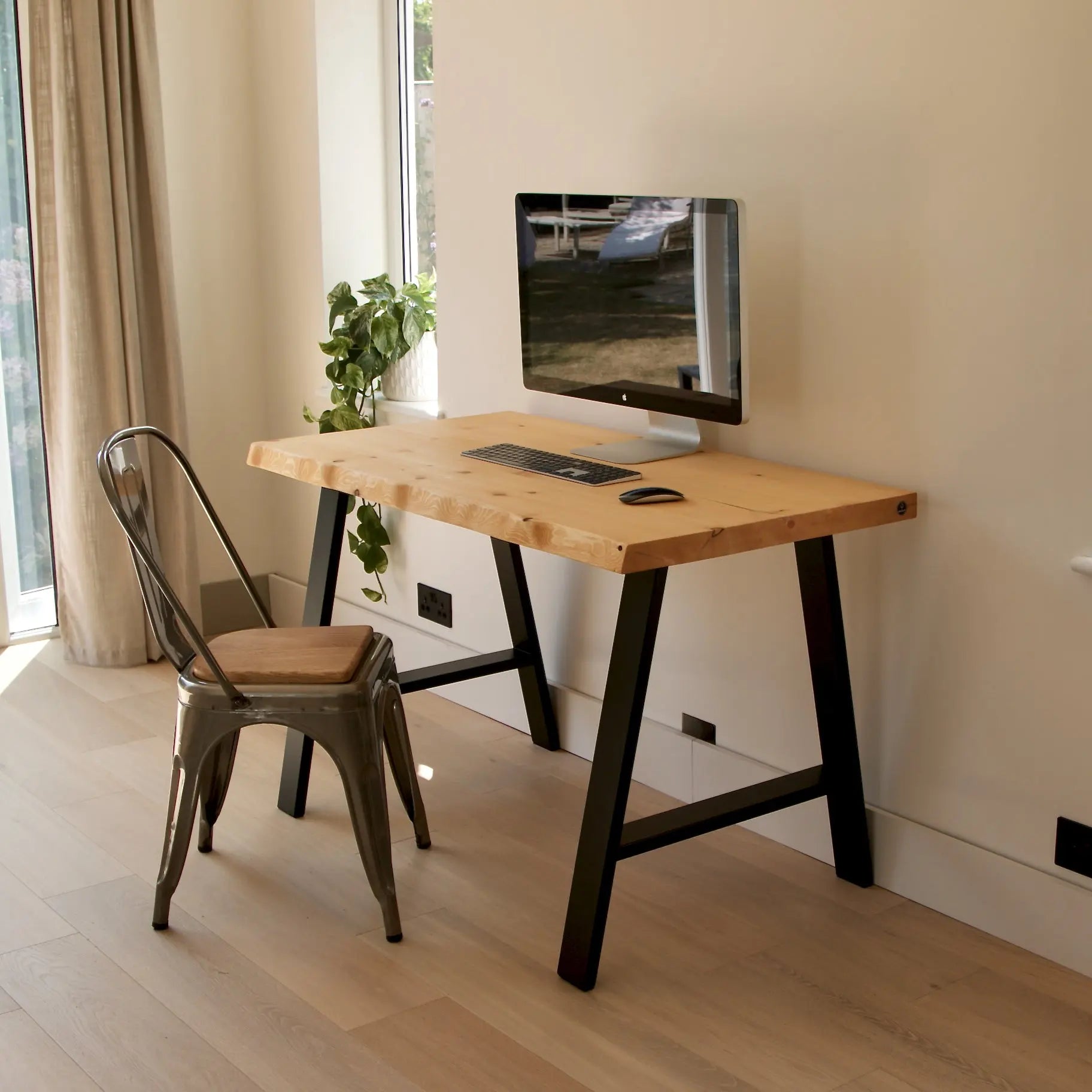 Wooden desk with black legs and a chair in a room with a television on the wall.