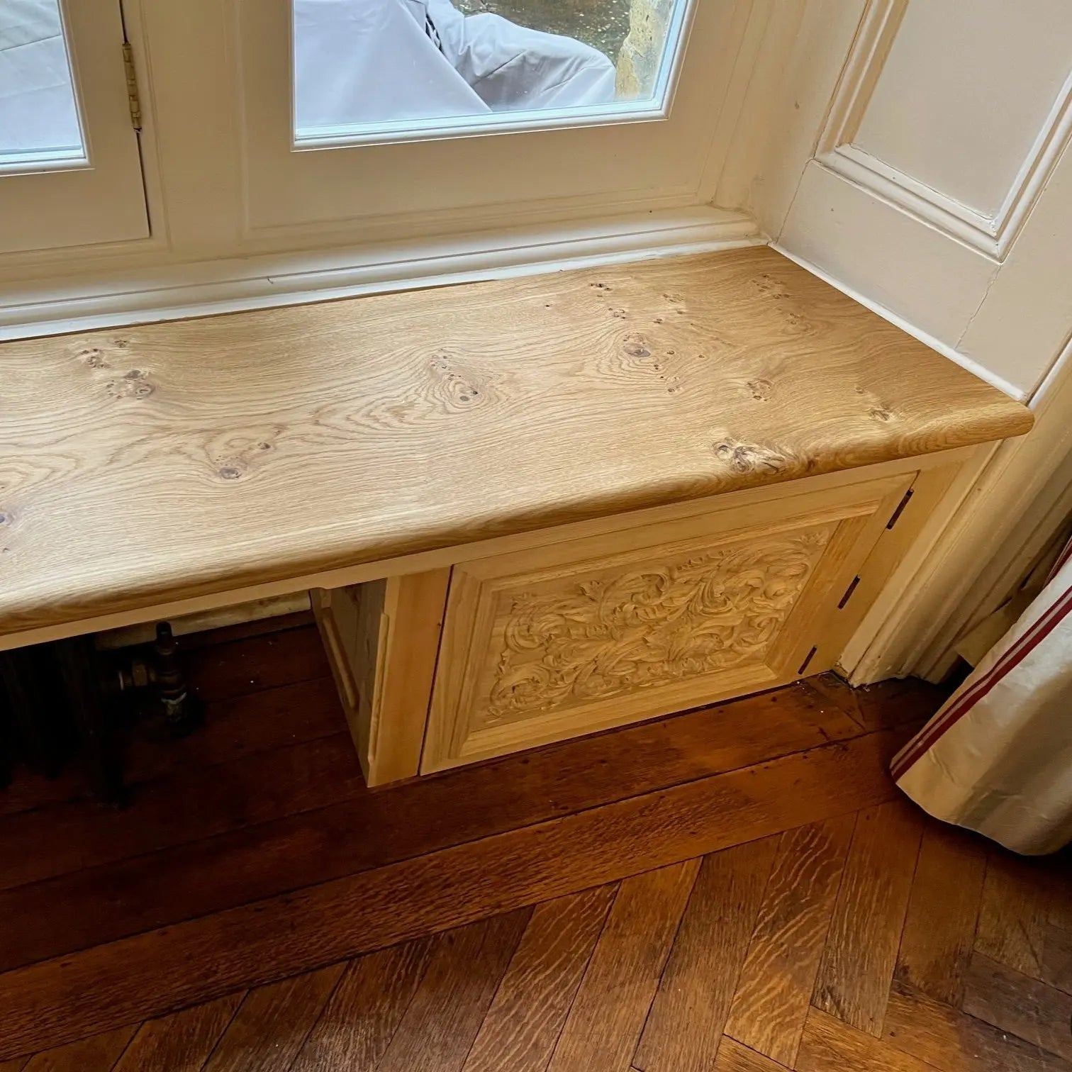 Wooden oak bench seat against a wall with a window above it on a wooden floor.